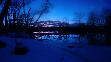 Lapso-De-Tiempo-De-Un-Pequeño-Puente-Rojo,-Arqueado,-De-Acero-Sobre-Un-Río-Al-Atardecer-Con-Reflejos-En-El-Agua-Y-Autos-Conduciendo-En-Un-Pequeño-Pueblo-De-América