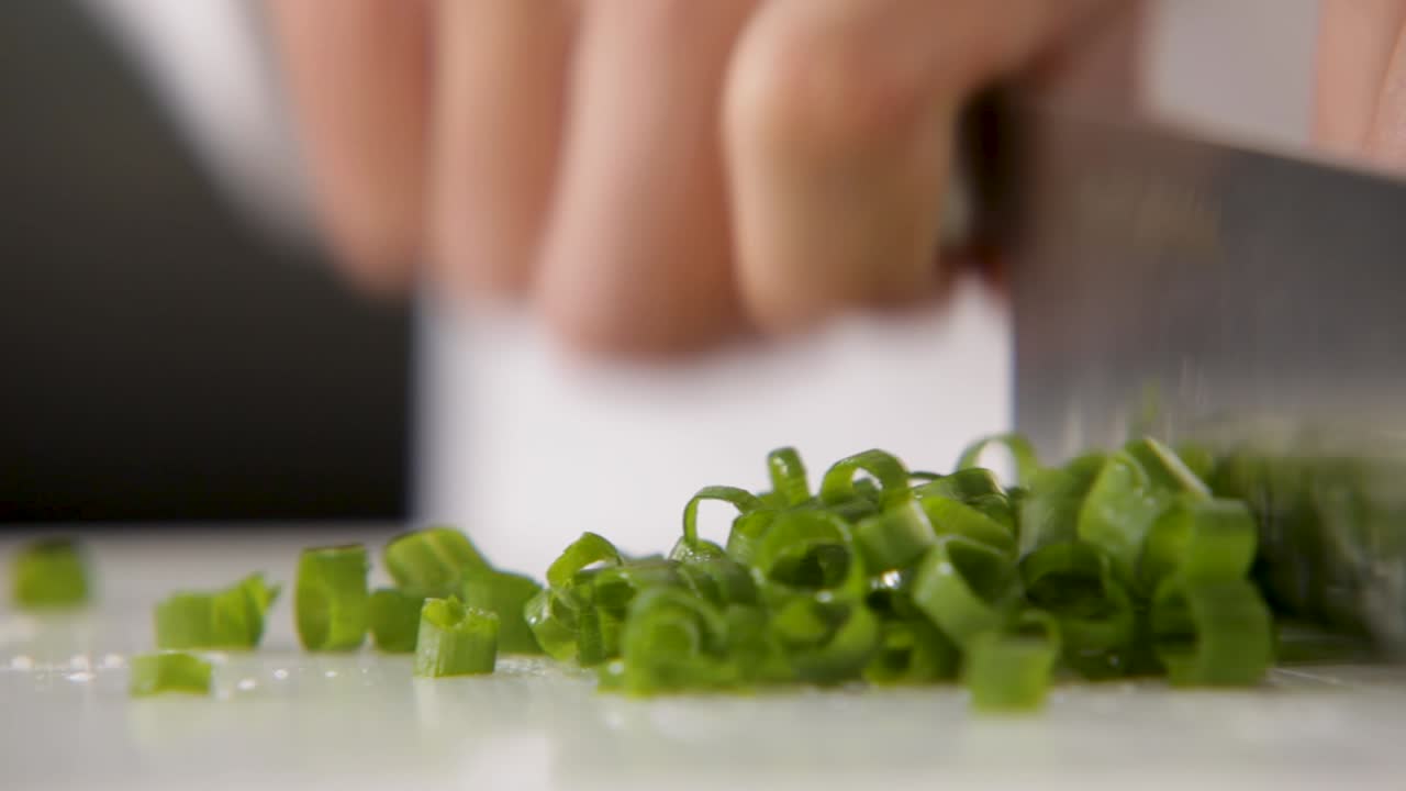 Close-up of green onions being finely chopped on a white cutting board, knife in motion