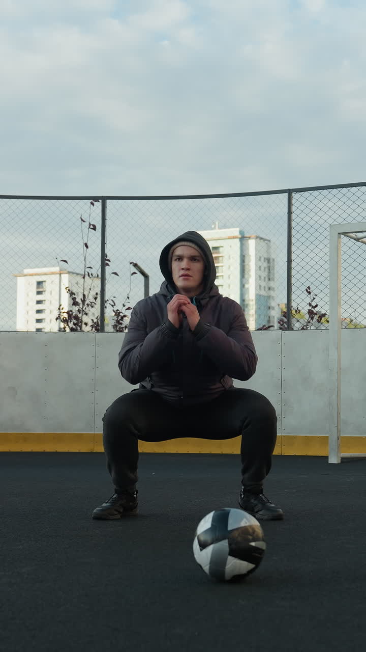 atleta realizando sentadillas con las manos juntas en preparación para el ejercicio en un campo deportivo al aire libre, una pelota de fútbol en primer plano, rodeado de oficinas urbanas de gran altura y postes de gol