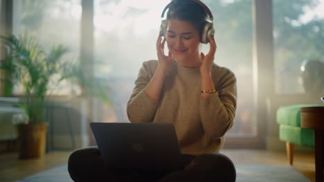young woman using laptop at home, does remote work, listens music through headphones. beautiful smiling girl sitting on the floor enjoys music, dances a bit, brainstorms creative project