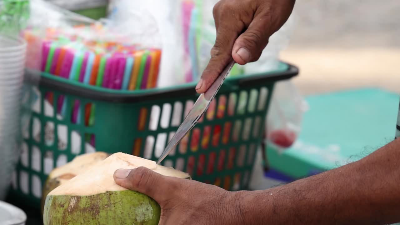 A vendor expertly slices open a coconut with a cleaver, readying it for serving at a vibrant market stall.