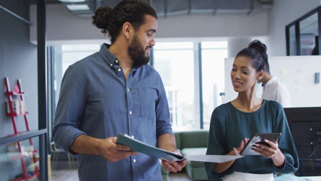 Two-diverse-happy-male-and-female-work-colleagues-walk-looking-at-tablet-and-documents-and-talking