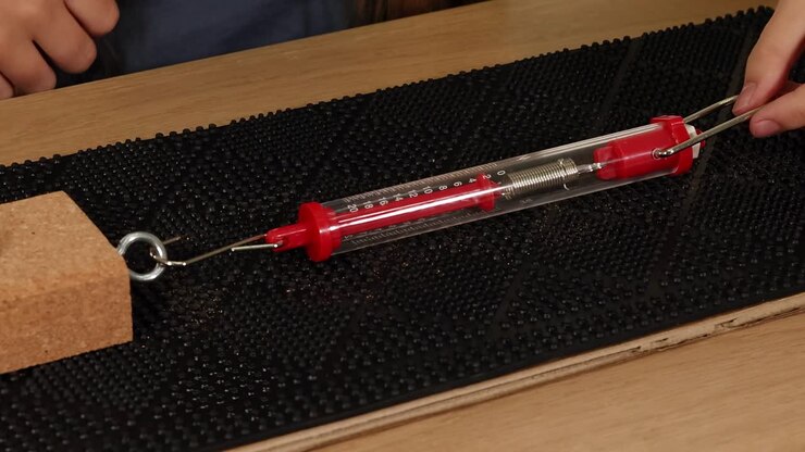 Hands demonstrate force measurement using a spring scale and metal weights on a textured surface.
