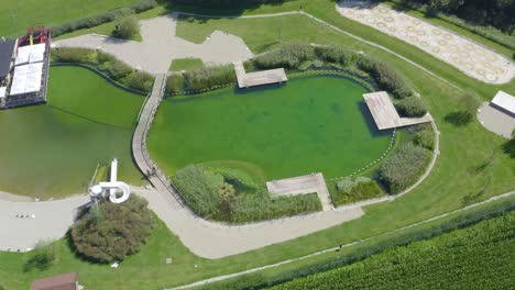 Pareja-De-Hombres-Y-Mujeres-Montando-En-Bicicleta-Alrededor-De-Un-Lago-Con-Un-Puente,-Tiro-De-Seguimiento-De-Vista-De-Pájaro-De-Drones-Aéreos,-Parque-De-Vodni-De-Eslovenia