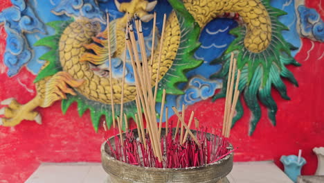 Burning-Incense-Sticks-With-Smoke-at-Hua-Hin-Thailand-Traditional-Buddhism-Temple-Against-Dragon-Depicted-on-the-Wall-Behind-the-Container