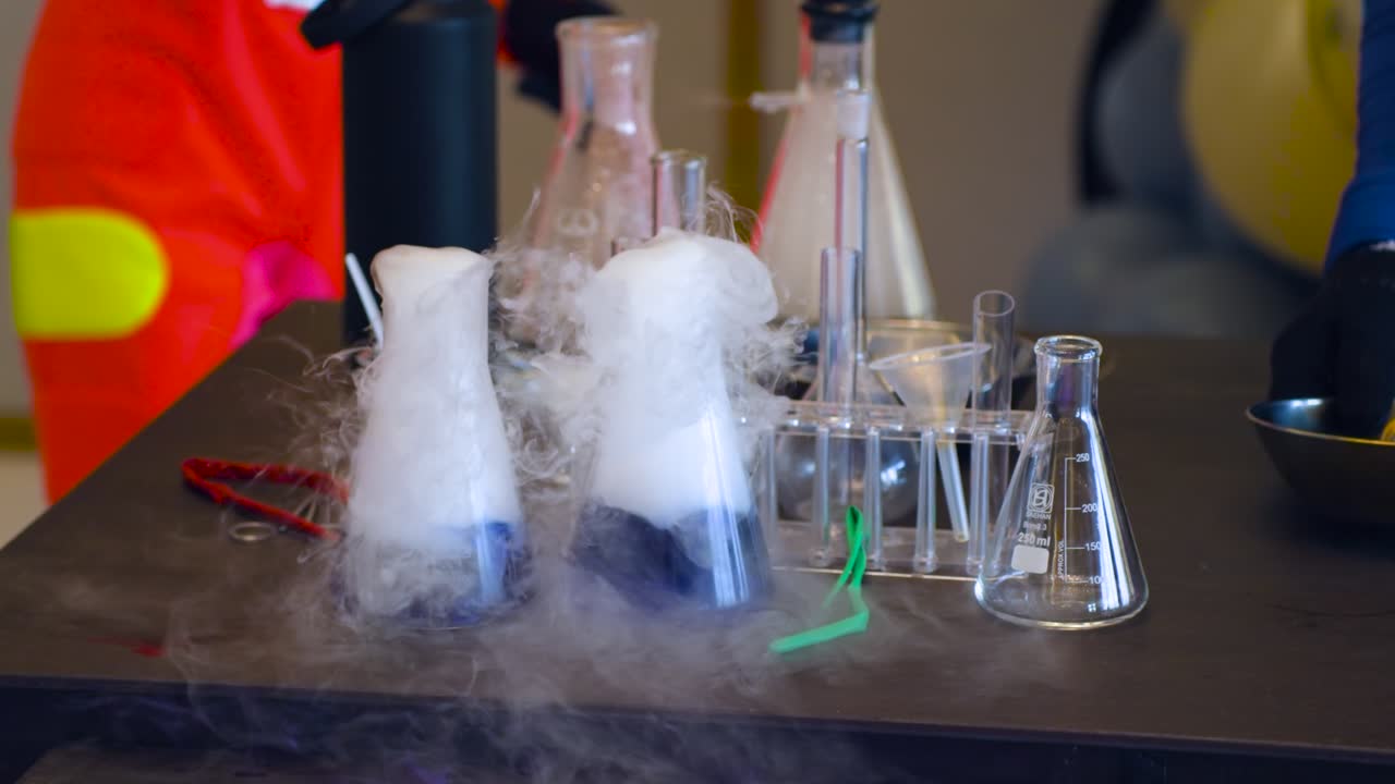 Science lab experiment with several Erlenmeyer flasks overflowing with thick white dry ice fog rising dramatically from colorful yellow and blue liquids surrounded by glassware and tools