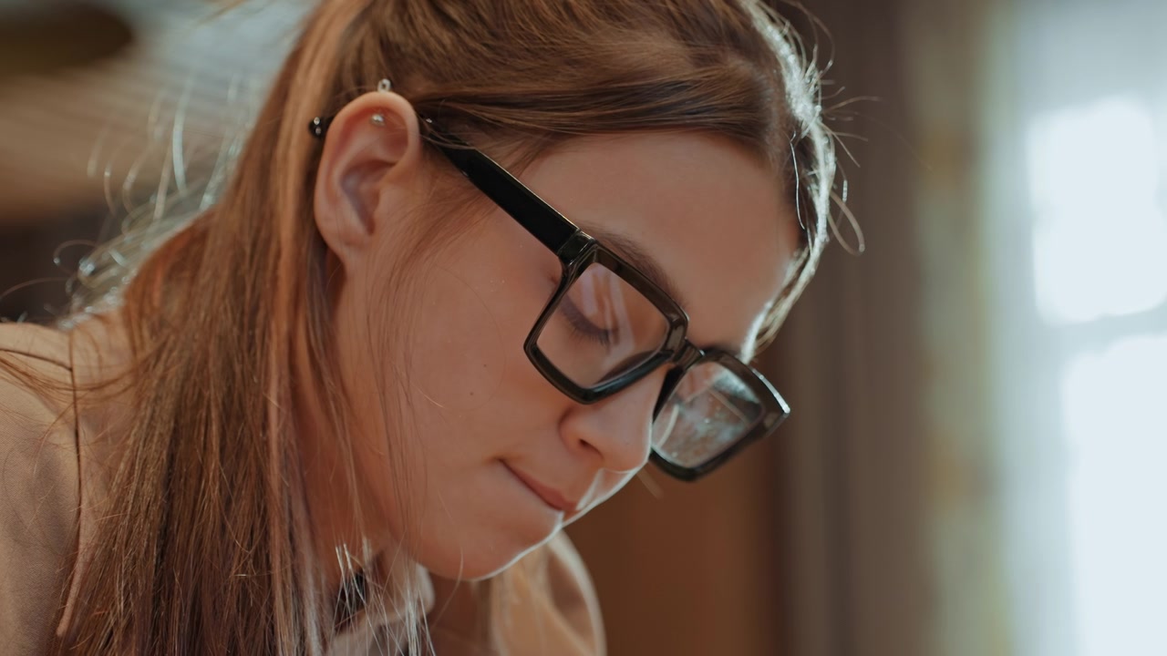 Donna caucasica impegnata in un'attività di apprendimento. Scena luminosa di una donna concentrata con gli occhiali che studia. Giovane donna serena con quaderno e penna immersa in uno studio riflessivo in luce calda.