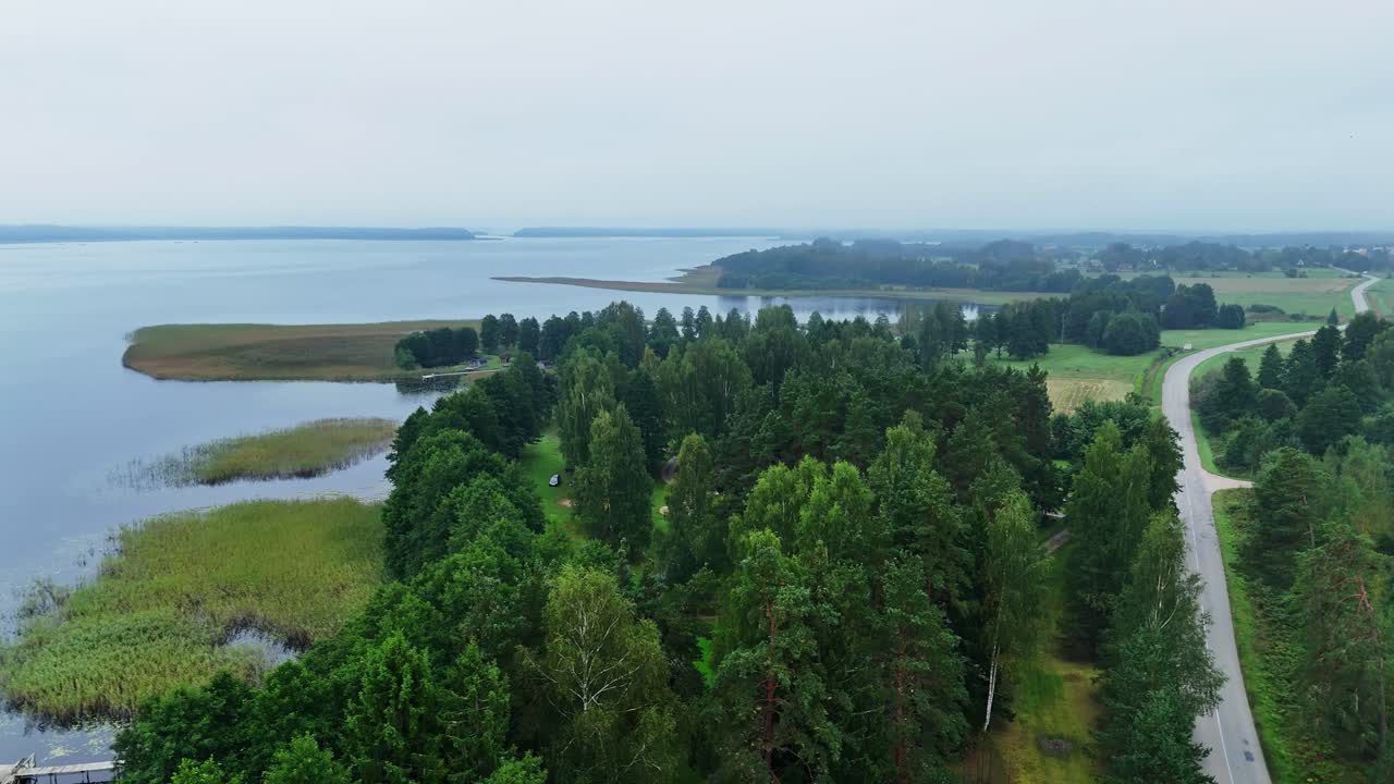 Aerial lateral shot of winding road, trees, and foggy lake shoreline with pier and fields