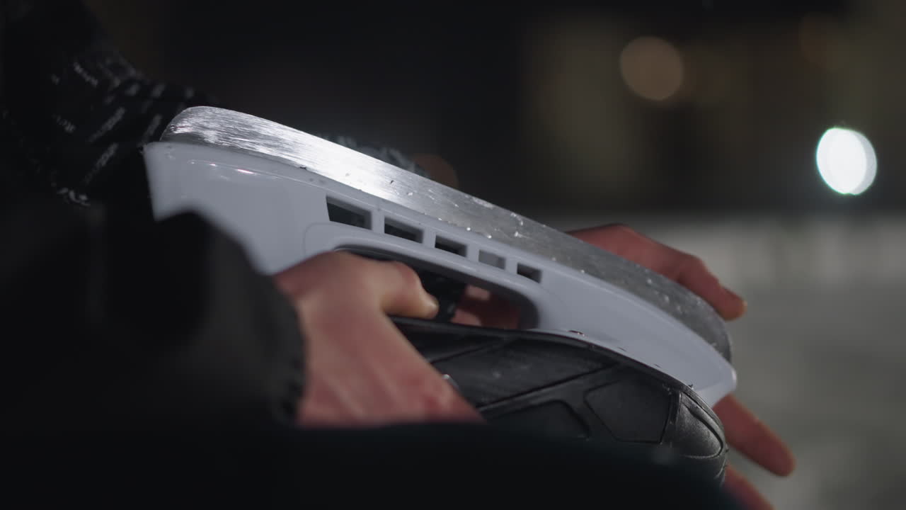 Close up of skater seated at night holding skate boot blade under soft bokeh lights with snowflakes falling gently in background, showcasing quiet preparation moment during cold winter evening
