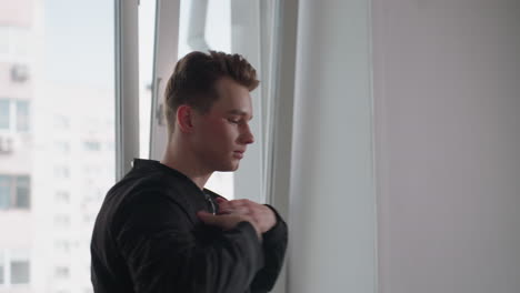 Young student stands by open window in bright urban apartment, lifts black jacket from arm and slips arms into sleeves while high rise building visible through window, preparing to head out