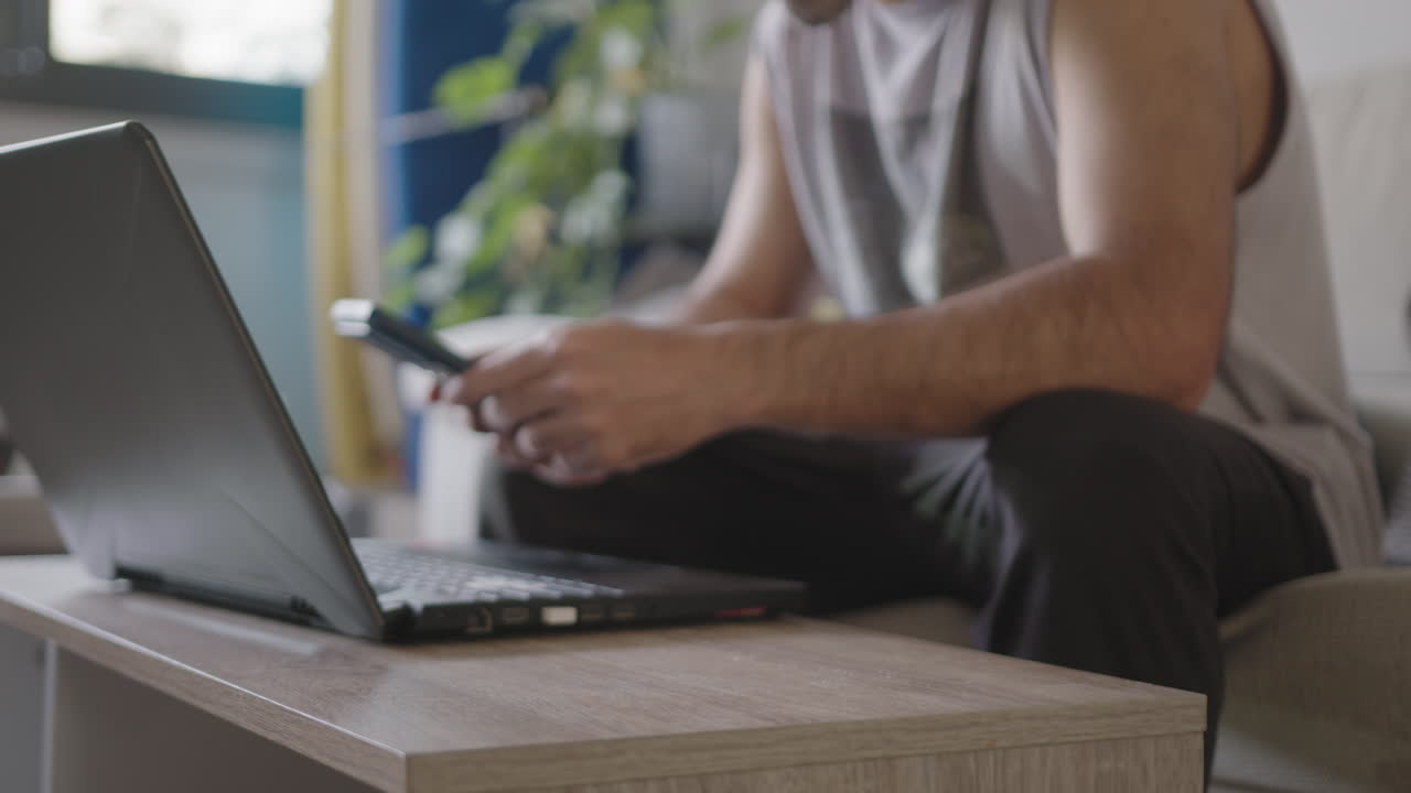Young adult male freelancer working from home in his living room using laptop, distracted from work with a phone urgent reminder pop up about is emails, sitting on the couch with casual clothes