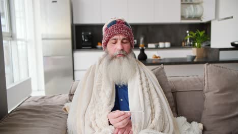 an elderly man with a gray beard is wrapped in a warm white blanket and checks his temperature with a thermometer. he sits in a hat and feels that he has a cold in a modern apartment