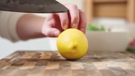 a male hand holding a knife cuts is cutting a fresh organic lemon into in a cross section half on a wooden cutting board inside a white minimalist kitchen