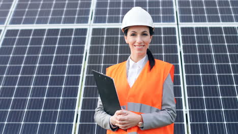 Portrait-of-young-female-engineer-in-special-uniform-and-helmet-holding-a-clipboard-near-solar-panels