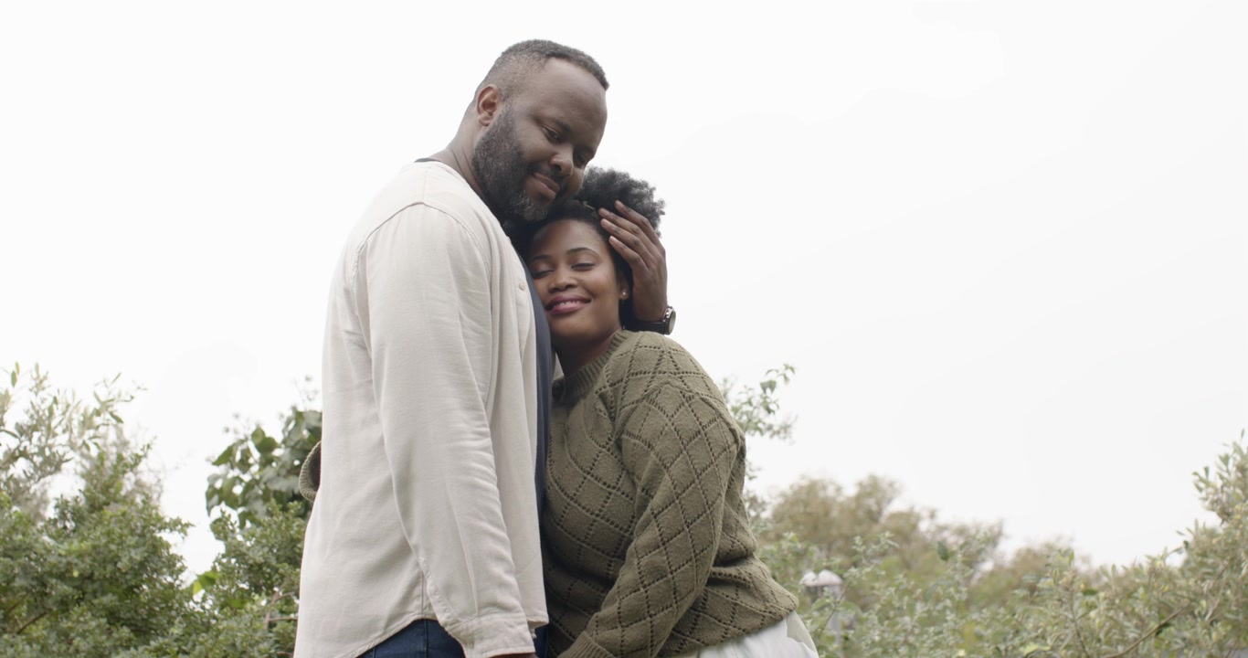 Couple holding hands at park, man lowering head, cupping hair, woman in olive sweater showing love