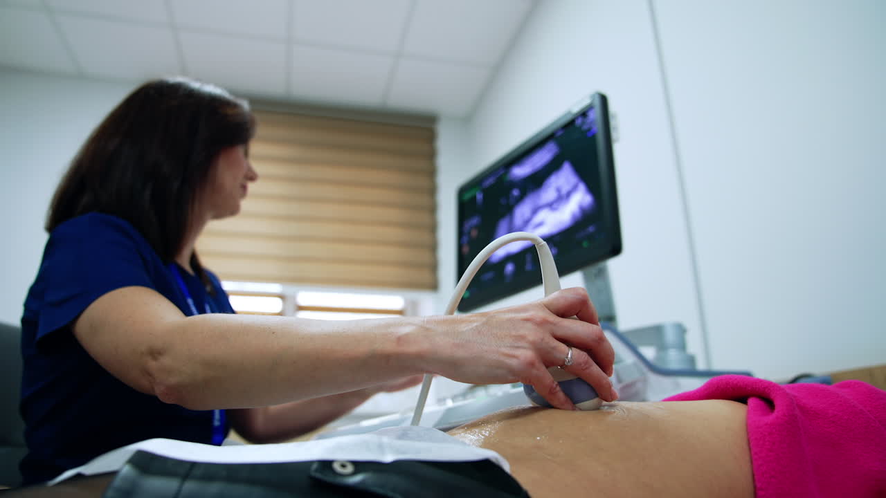 Female brunette doctor check the pregnancy of a patient at ultrasonic equipment. Specialist holds the transducer on woman's belly. Low angle view.