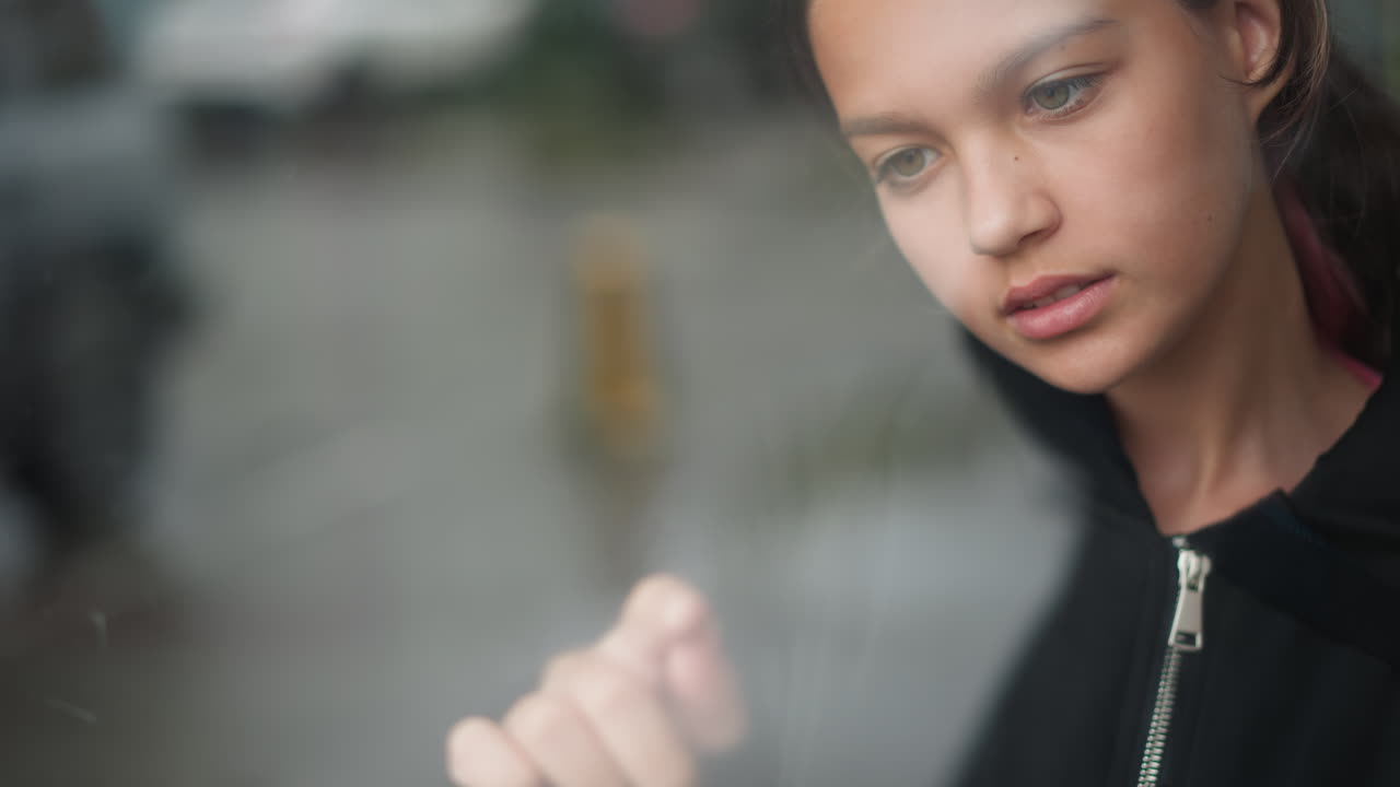 Молодая женщина наблюдает за дождливой сценой. Женщина смотрит на капли на стекле с размытым городским фоном. Молодая женщина у забрызганного дождем окна созерцает далекий трафик и отражения городского пейзажа