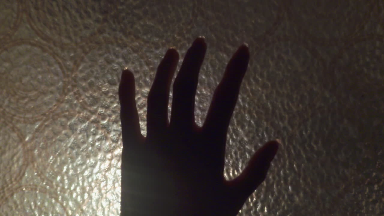 A cinematic tracking shot following a woman's hand as it slides across a frosted textured glass window pane during sunset in a cozy Budapest Hungary apartment interior during the year 2026