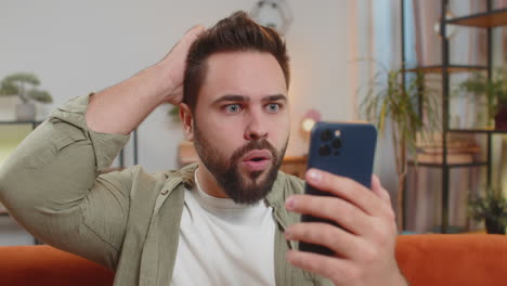 surprised caucasian brunette man using smartphone winning lottery sitting on sofa couch at home