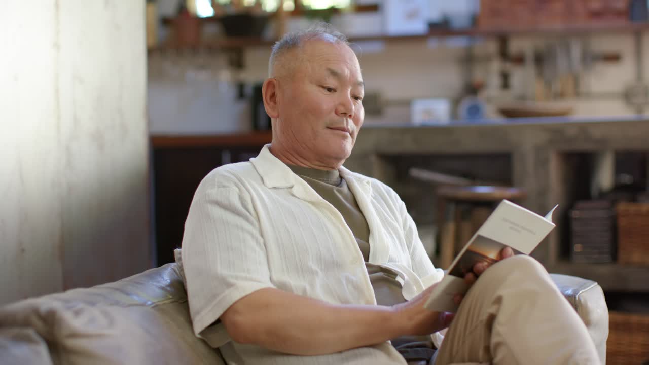 自宅のソファに座ったアジア系の高齢男性が、柔らかな自然光の下で文庫本を読み、ページをめくっている。