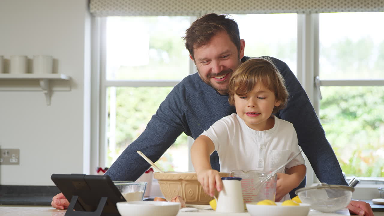 padre e hijo en pijama horneando en la cocina en casa siguiendo la receta en una tableta digital