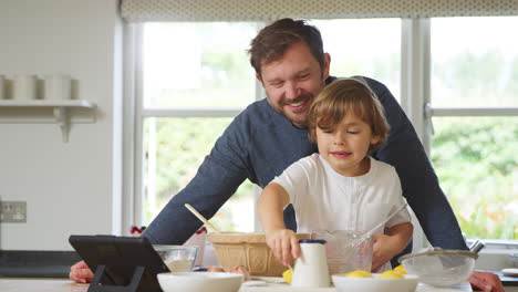 Padre-E-Hijo-En-Pijama-Horneando-En-La-Cocina-De-Casa-Siguiendo-La-Receta-En-Una-Tableta-Digital