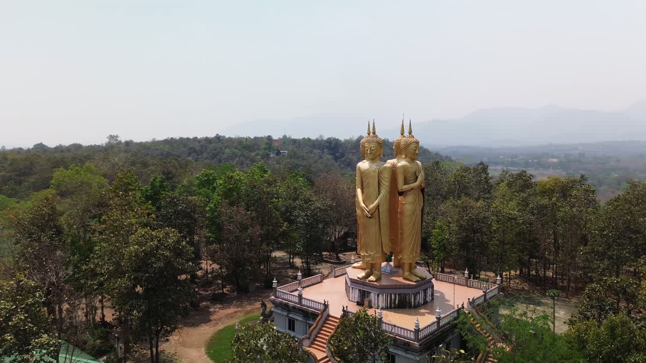 Wide aerial orbit of gold Buddha statue near Chiang Mai, Thailand surrounded by trees and hills with prayer hands on pedestal
