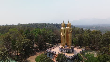 wide aerial orbit of gold buddha statue near chiang mai, thailand surrounded by trees and hills with prayer hands on pedestal