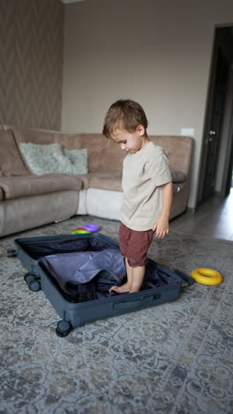 cute caucasian boy is hiding in the big suitcase. kid stands up and goes to pick up his clothes scattered by the room. vertical video.