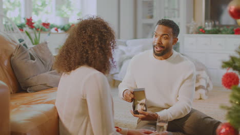 Loving-Couple-Drinking-Champagne-As-Man-Opens-Gift-Of-Wristwatch-Around-Christmas-Tree-At-Home