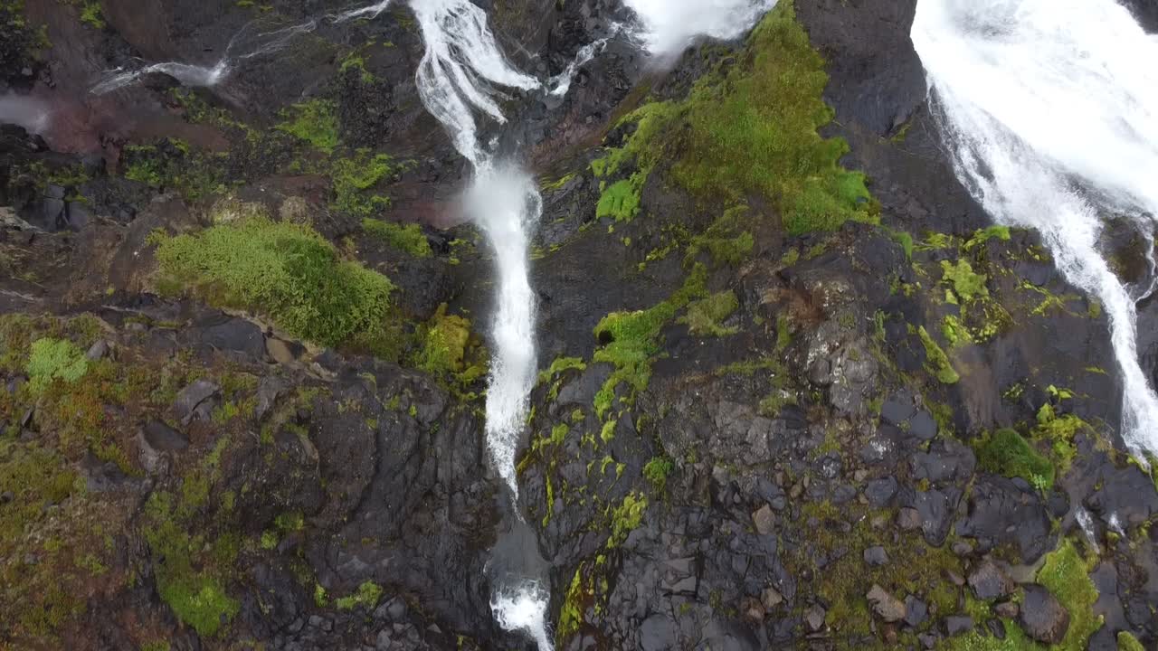 toma aérea de arriba hacia abajo de una idílica cascada en islandia que fluye por montañas rocosas y cubiertas de musgo