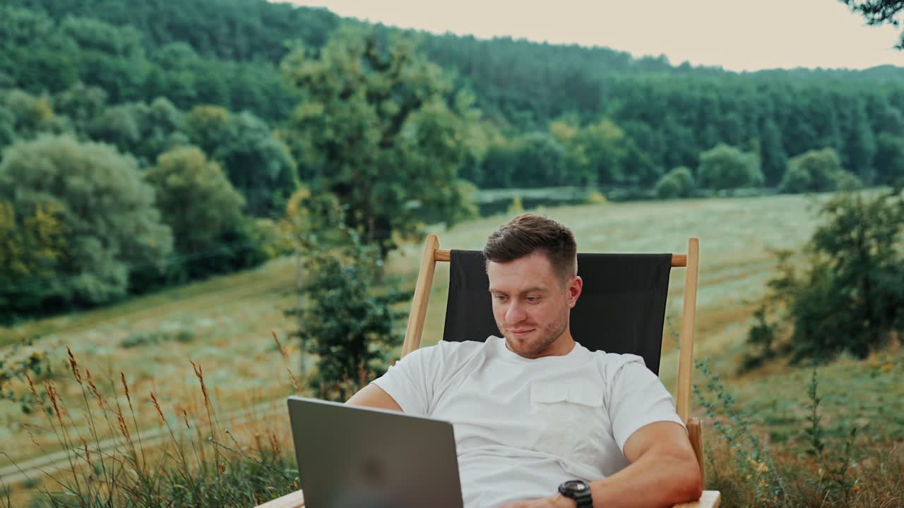 Work in the nature far away from the office. Relaxed happy freelancing man working on his laptop sitting in a folding chair.