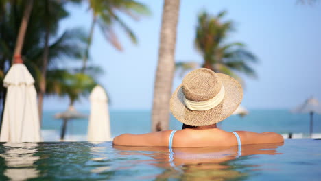 a woman, with her back to the camera, in a straw sun hat leans along a pool edge looking out at the ocean view