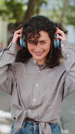 Portrait-of-happy-smiling-woman-listening-music-taking-off-headphones-looking-at-camera-outdoors