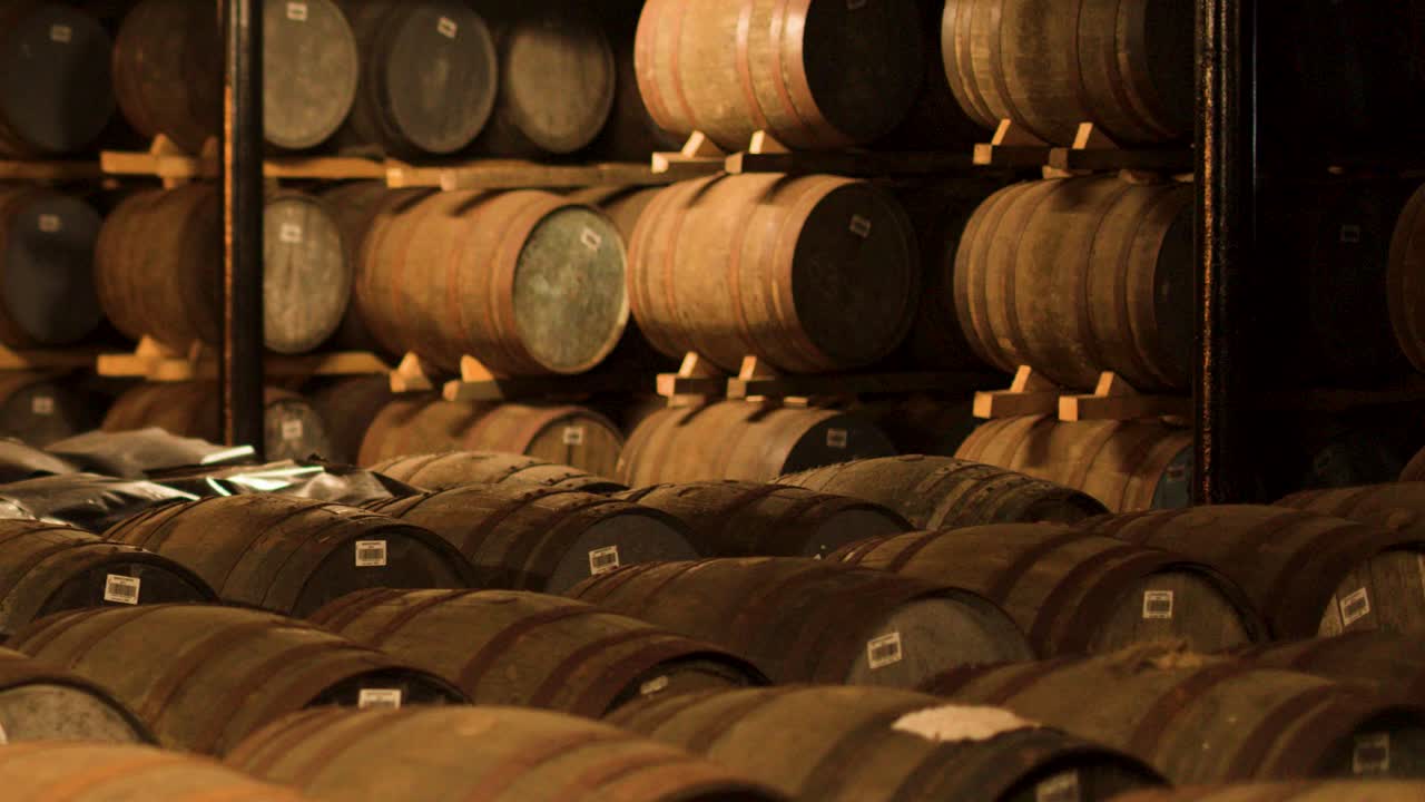 Oak barrels aging whisky in a dim, rustic warehouse with slow lateral camera movement