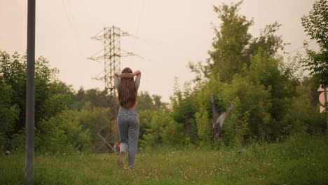 joven caminando por un camino cubierto de hierba con la mano en la cabeza en postura contemplativa, rodeada de exuberante vegetación y líneas eléctricas lejanas, el fondo muestra luz roja borrosa y un coche que pasa