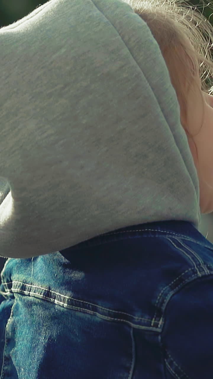 CU, slow motion: pretty happy little girl in jacket with grey textile hood has fun jumping and looking upwards on sunny day on blurred background close view