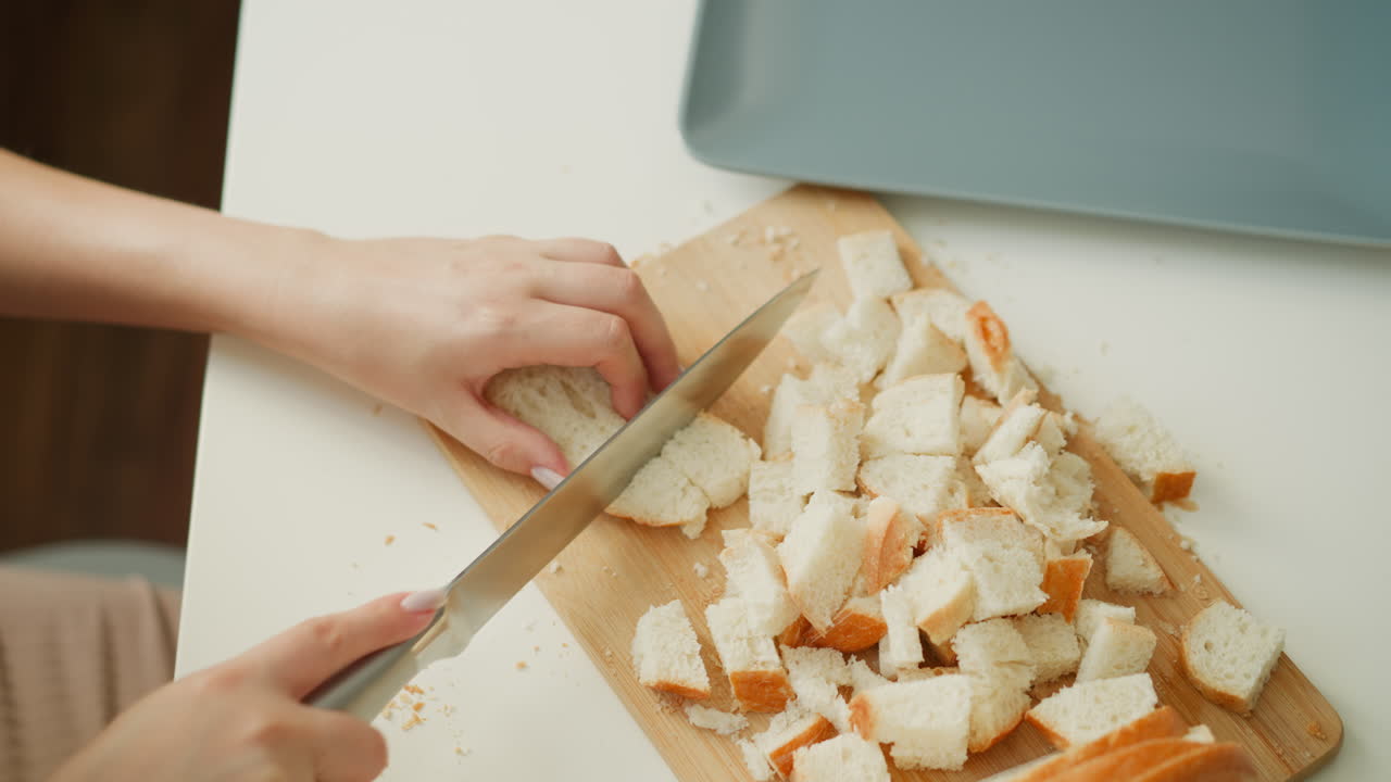 High angle view of female hand chopping cake slices into smaller pieces on wooden board over white table under soft window light, crumbs scattered, showcasing precise culinary preparation