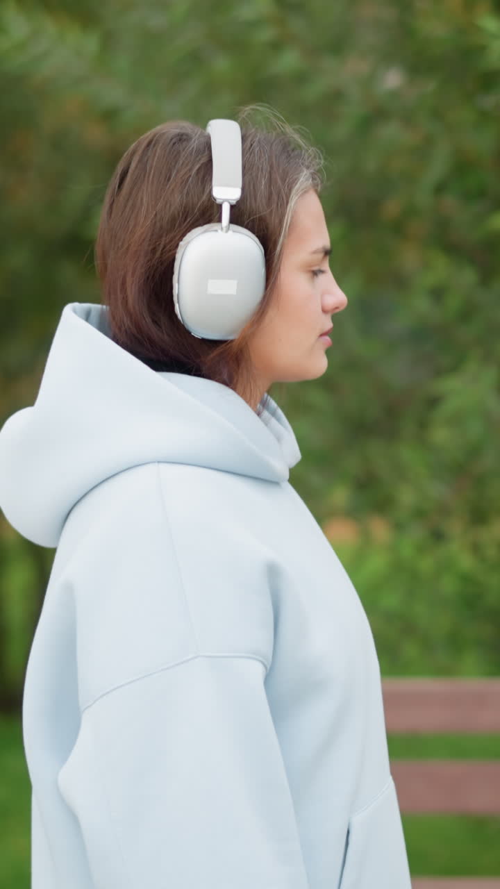 Giovane donna che cammina attraverso un giardino, circondata da fiori con una panchina di cemento sullo sfondo, che indossa cuffie bianche e una felpa casual con cappuccio, ideale per scene di benessere, natura o relax all'aperto.