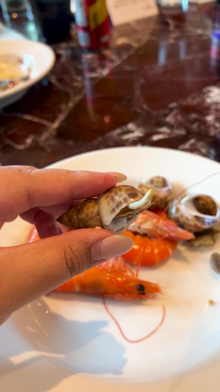 A person holds a cooked sea snail over a plate of shrimp at a restaurant