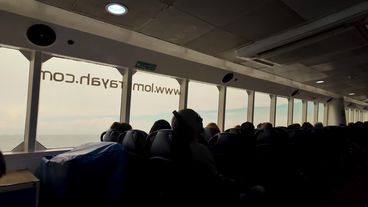 Silhouetted passengers seated inside a speedboat ferry during a daytime journey. Natural light illuminates through large windows, creating a serene travel atmosphere on the water.