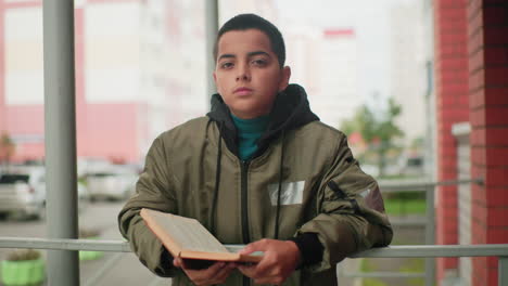 little boy in green jacket holds open book with reflective expression, standing against brick wall with soft blurred urban background and parked cars, creating a thoughtful atmosphere