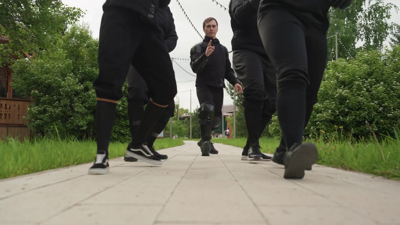 Close up of three people walking forward on paved path surrounded by greenery. people wear black clothing and protective gear with their legs visible as they move through outdoor environment