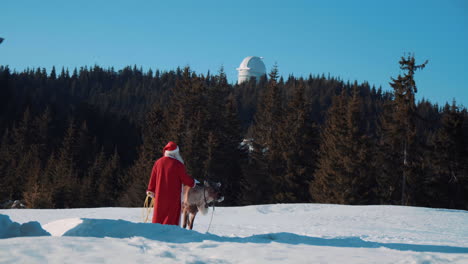 santa claus and deer rudolph stand on a snowy meadow and look at a telescope