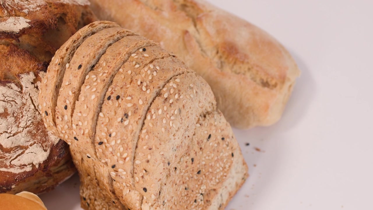Close-up panning shot of sourdough, French baguette, and sliced multigrain bread on white background.