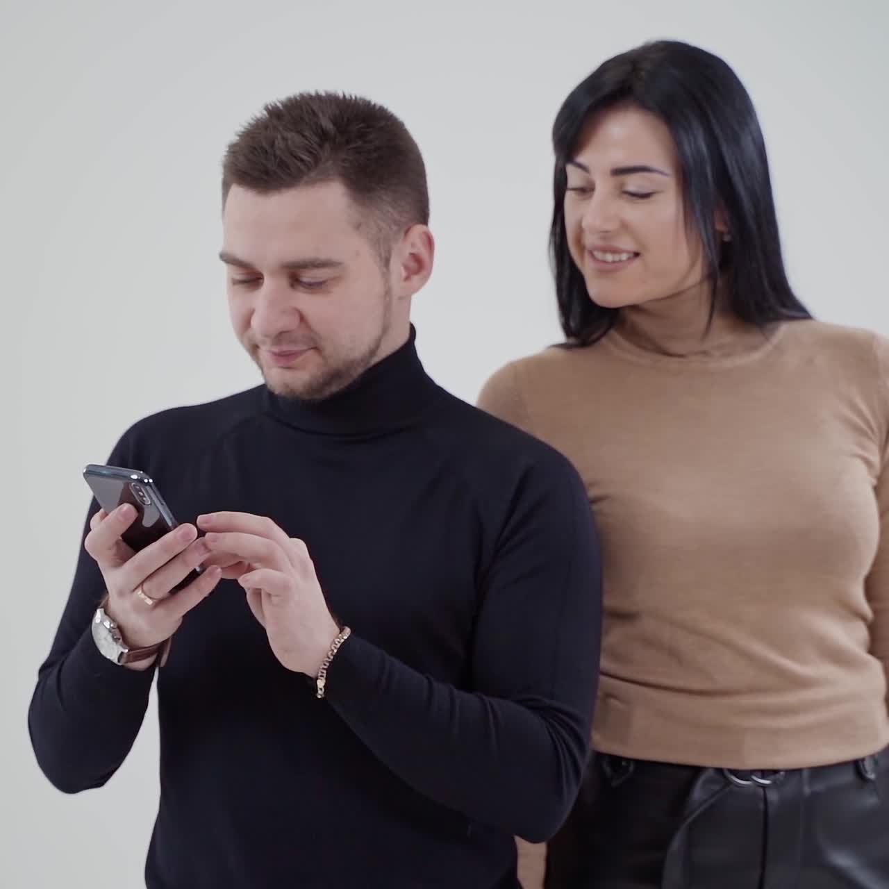 Young happy couple looking at a phone in studio. Man in black sweater holds a cell phone and a woman standing behind him smiling. Slow motion.