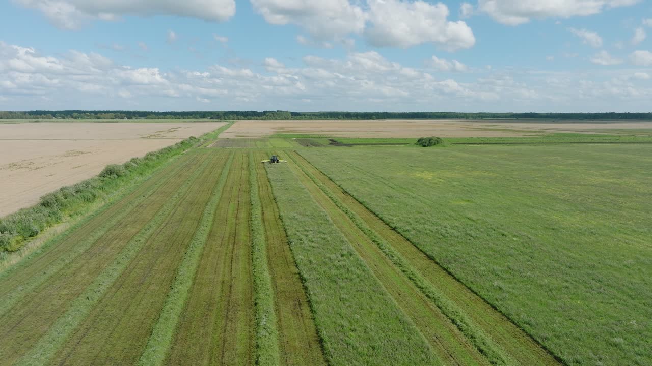 Aerial establishing view of a tractor mowing a fresh green grass field, a farmer in a modern tractor preparing food for farm animals, sunny summer day, wide drone shot moving forward low