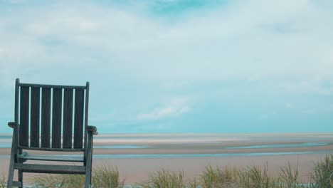 Weite-Aufnahme-Eines-Einzelnen-Holzstuhls,-Der-Bei-Ebbe-Auf-Einen-Strand-Blickt,-Wo-Sich-Wasserschlangen-Bis-Zum-Horizont-Erstrecken,-Wo-Sich-Ein-Gemeiner-Sturm-Zusammenbraut