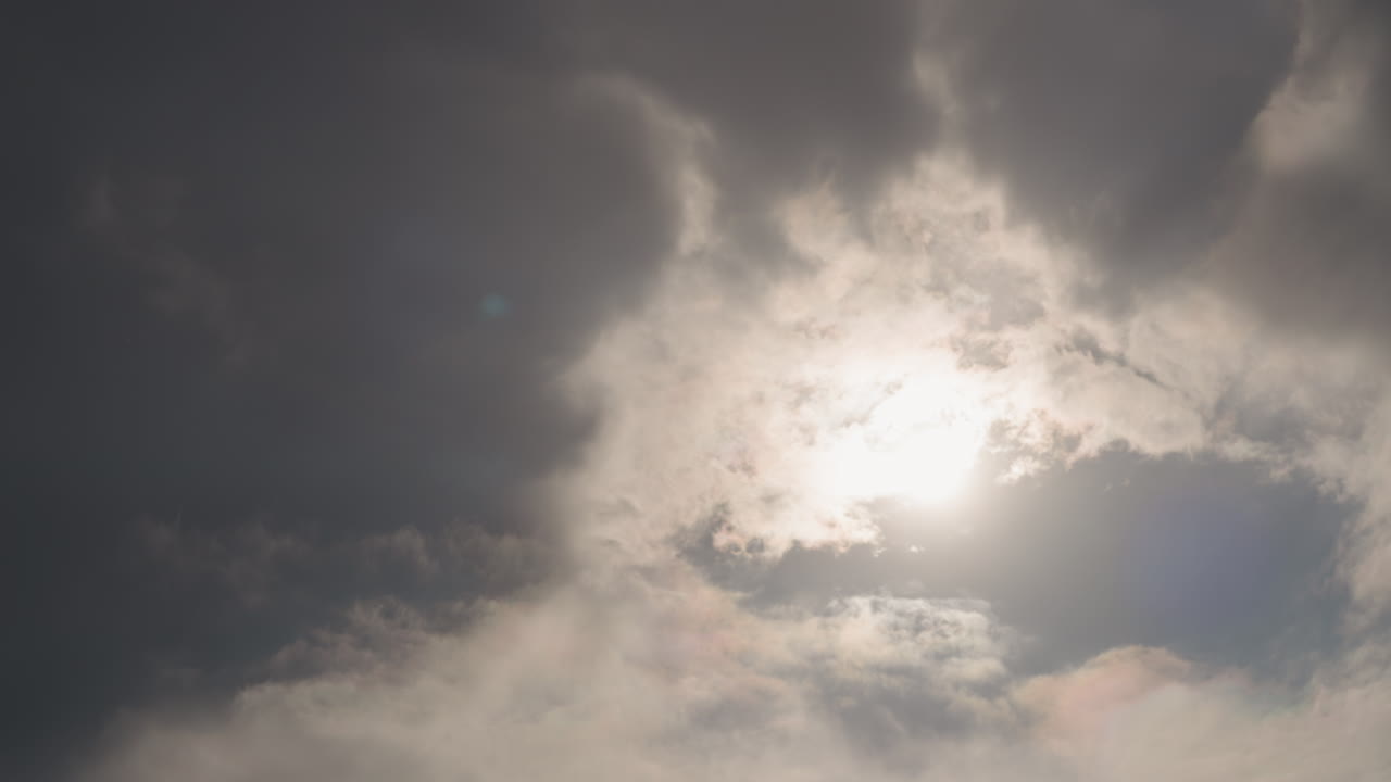 sol que se filtra entre nubes de tormenta, capas de cirros y cúmulos retroiluminadas de forma espectacular, rayos luminosos y sutil destello de lente, atmósfera sombría con toques de lluvia y llovizna, toma cinematográfica amplia ideal