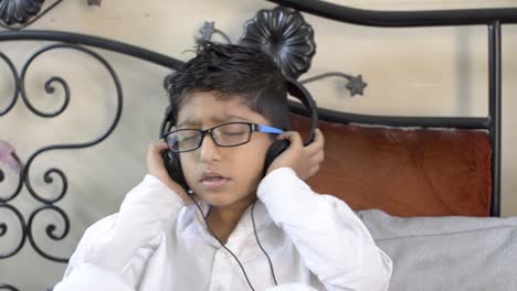 little indian asian caucasian boy wearning glasses spectacles listening enjoying music on headphone closing eyes and hands on ear sitting in living room side view